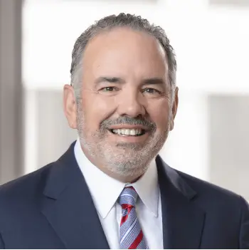Smiling middle-aged man in a navy suit and red striped tie, standing in a bright office