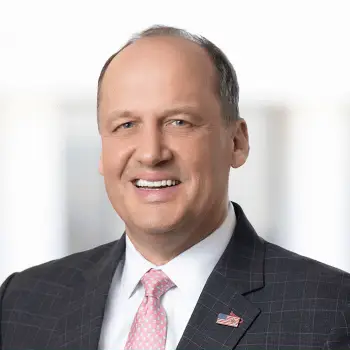 Smiling businessman in a dark plaid suit with a pink tie and an American flag lapel pin.