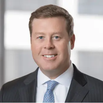 Smiling man in a dark pinstripe suit and light blue tie, standing in a bright office setting.