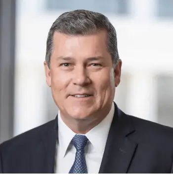 Portrait of a smiling middle-aged man in a dark suit and blue tie, standing by a window.