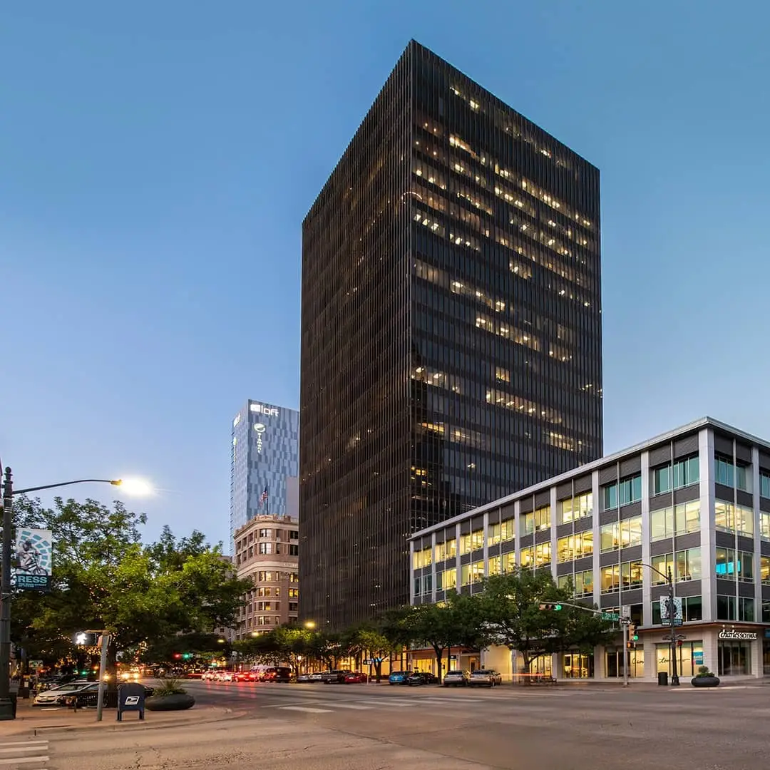 Tall, dark skyscraper stands illuminated at dusk on a city street, amongst other buildings and trees.