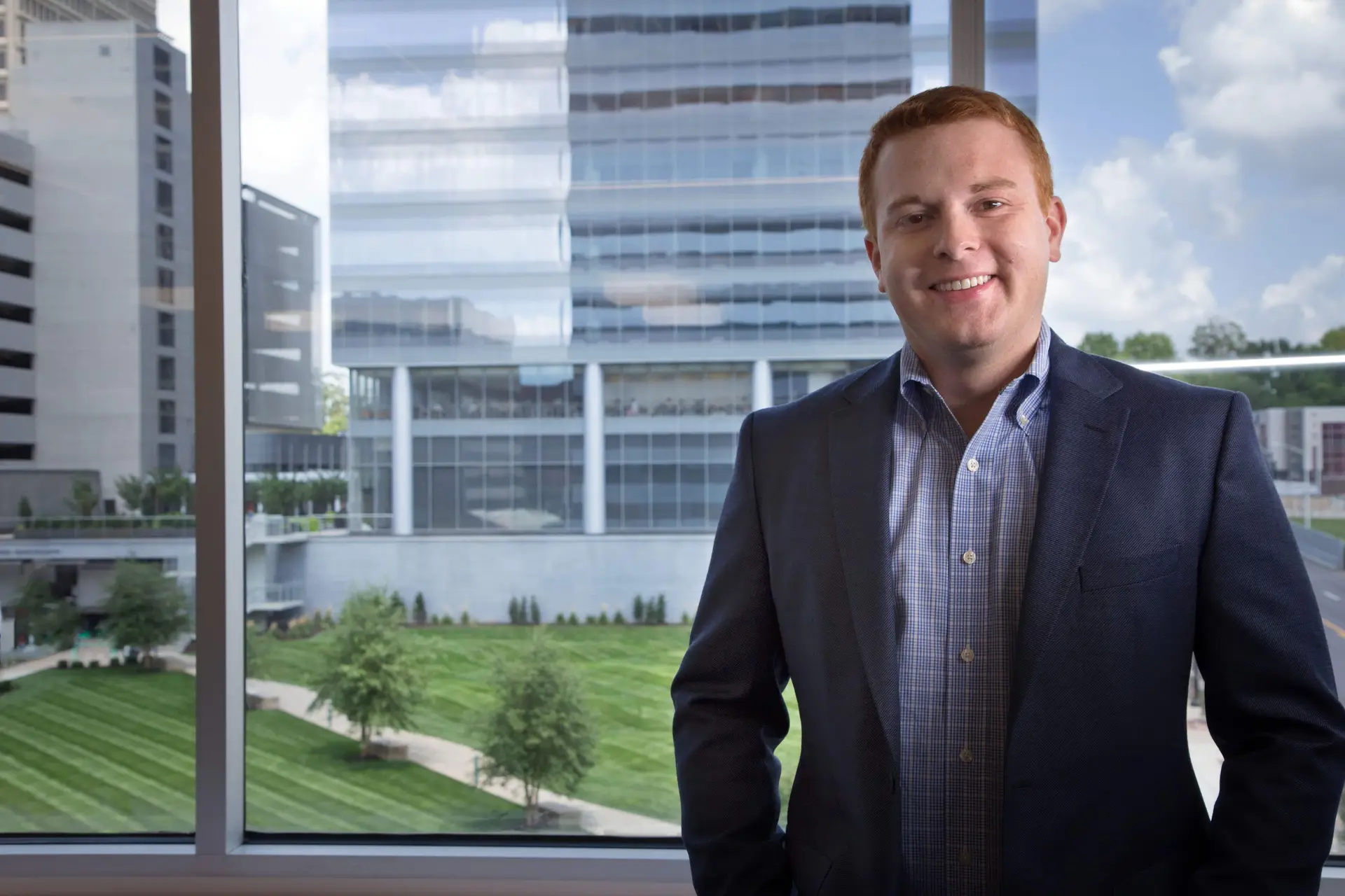 Smiling man in a navy blazer and checkered shirt standing by a large window with glass office buildings in the background.