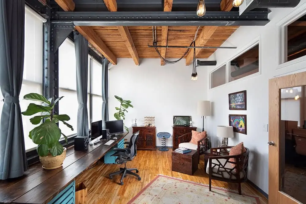 Bright loft office with a long wooden desk by tall windows, with plants and a seating area beneath exposed beams.
