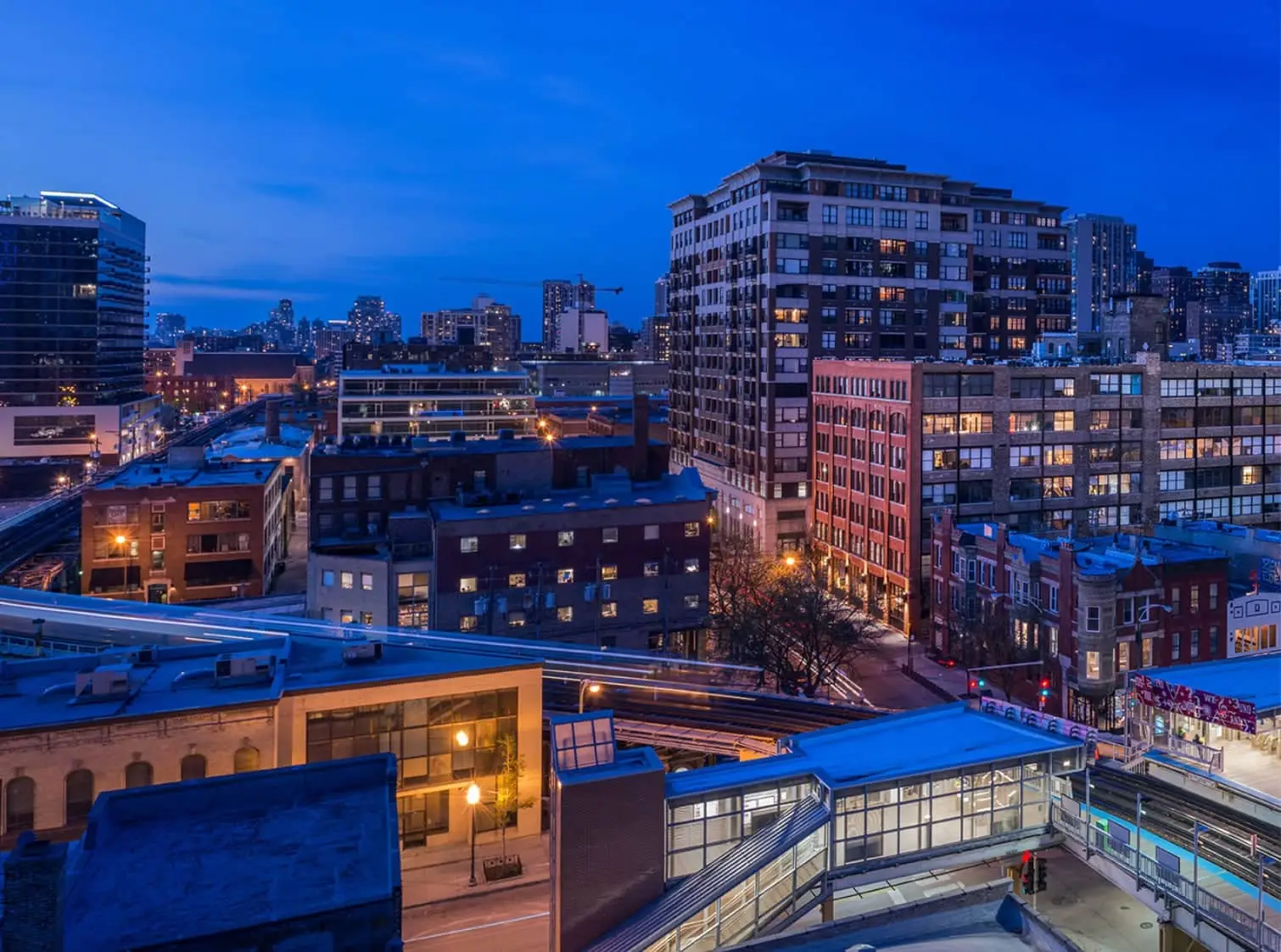 City skyline at dusk with apartment buildings, elevated tracks, and streetlights.