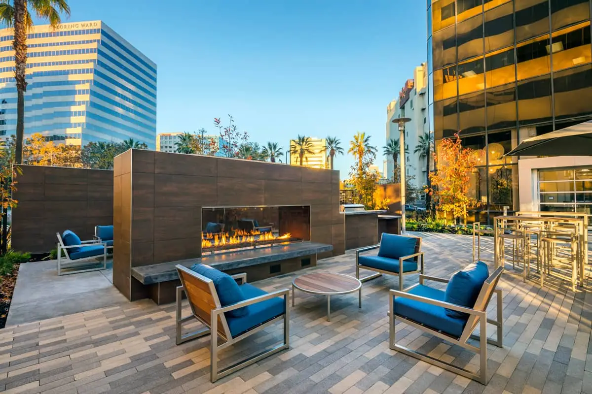 Outdoor courtyard with a long fireplace, blue-cushioned seating, and metal chairs in a modern urban plaza.