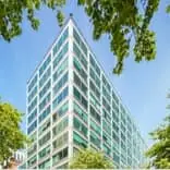 Modern glass office building with a grid of windows, framed by leafy trees under a clear blue sky.