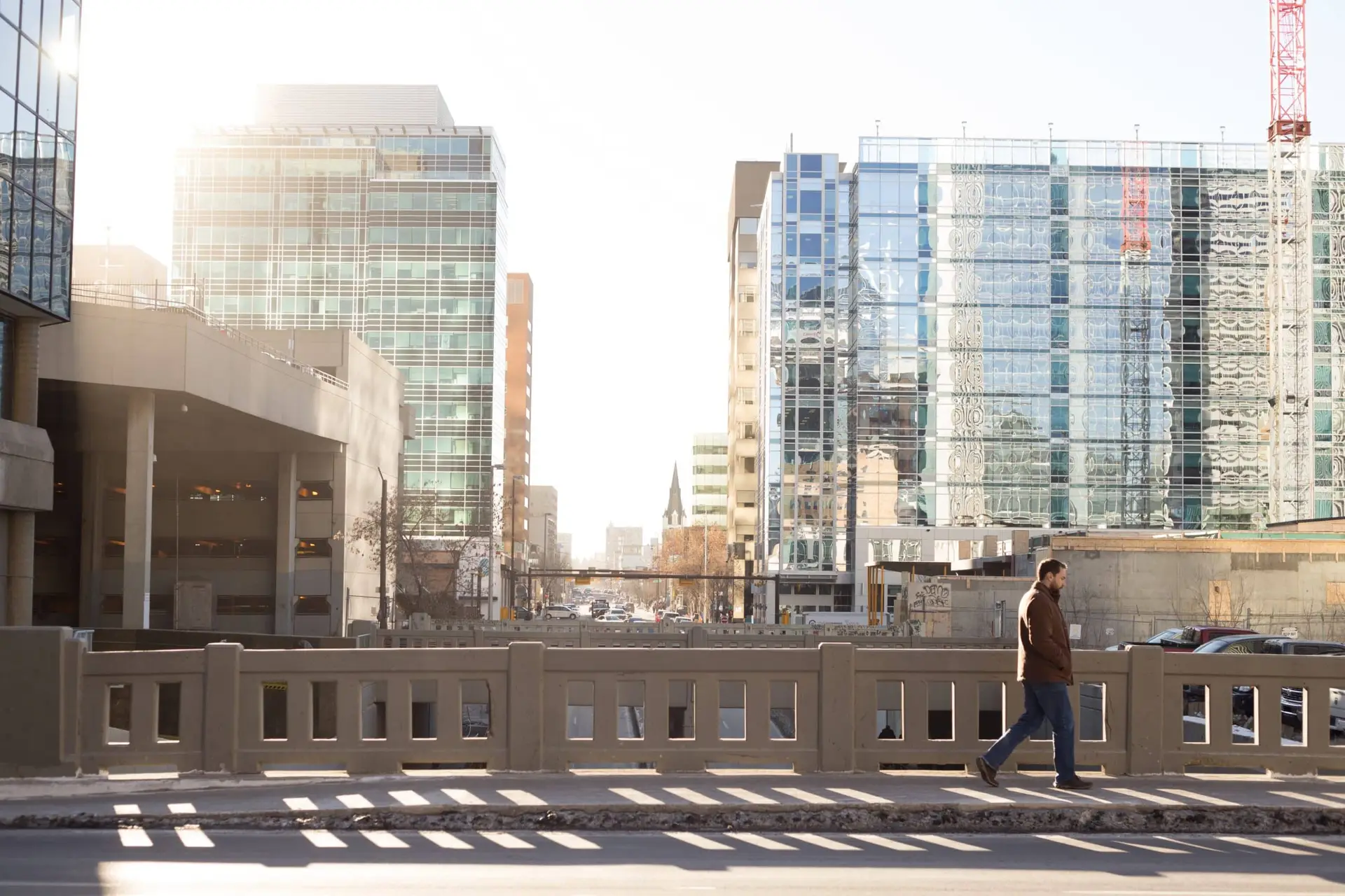 Sunlit city street with modern glass office buildings and a lone man walking along a concrete barrier.