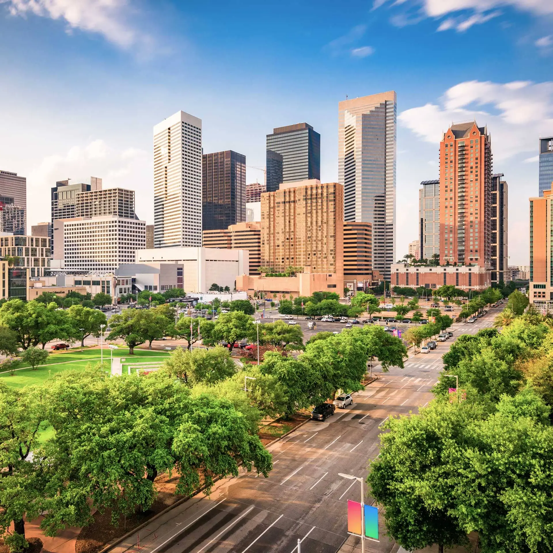 Skyline of modern skyscrapers rising above a green park and busy city street.