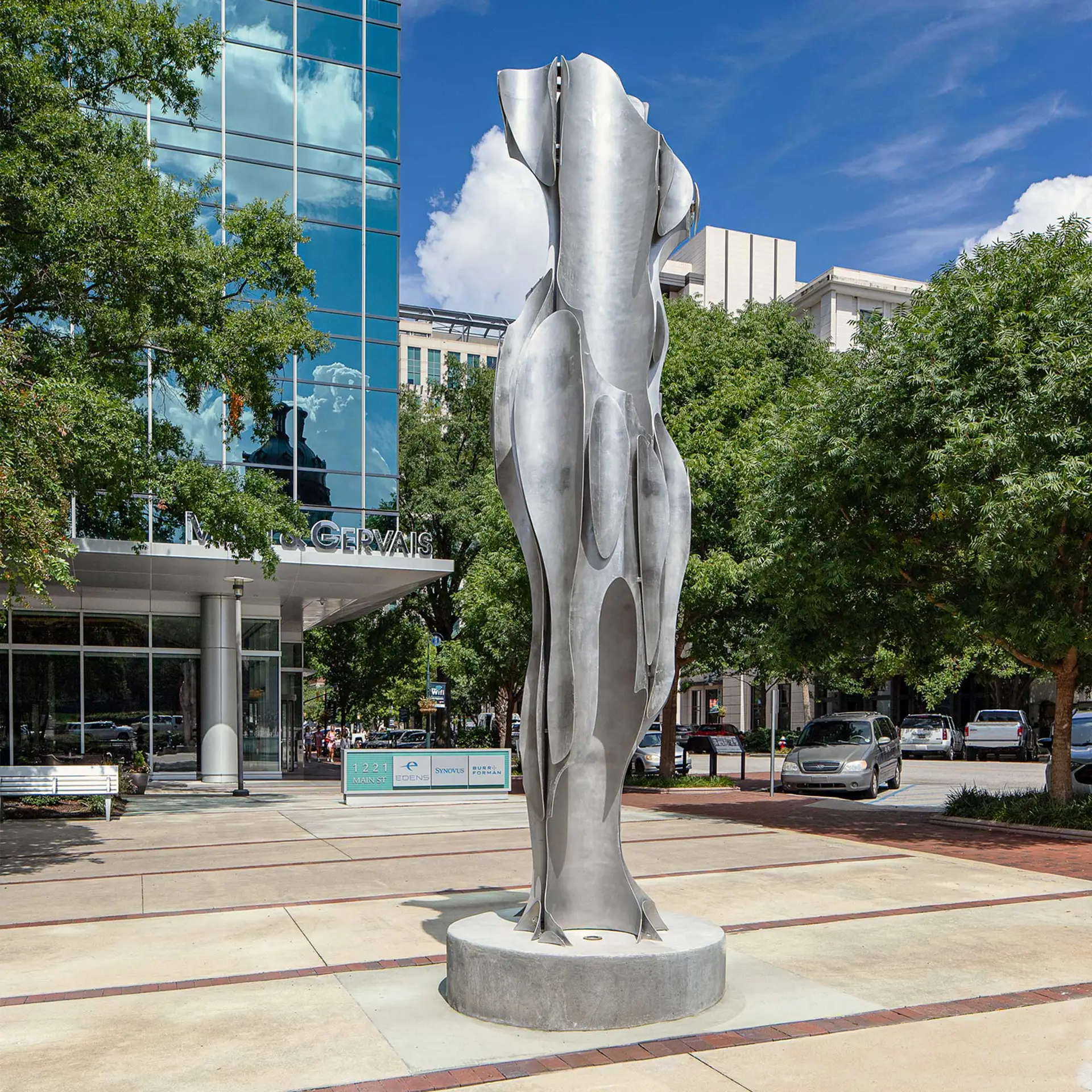 Abstract metallic sculpture in front of a glass office building with green trees and a blue sky.