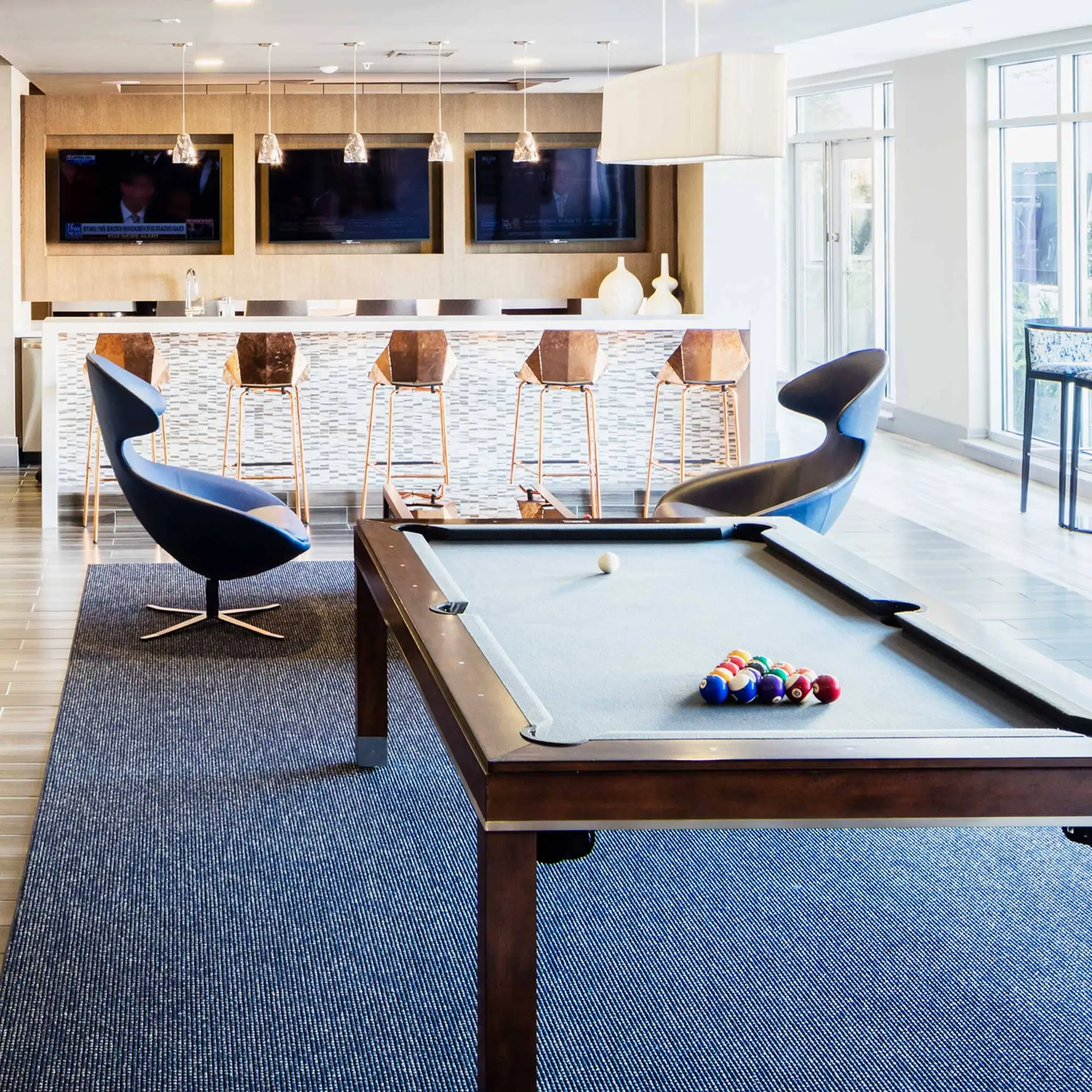 Modern lounge featuring a pool table, blue curved chairs, and a bar with copper stools.