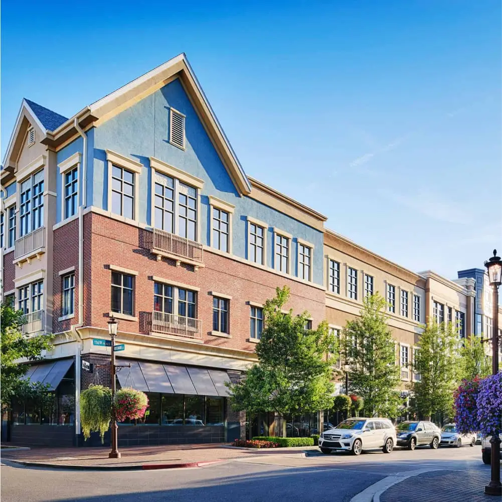Multi-story brick building sits on a corner, showcasing retail space and parked cars. The street is quiet, under a bright blue sky.