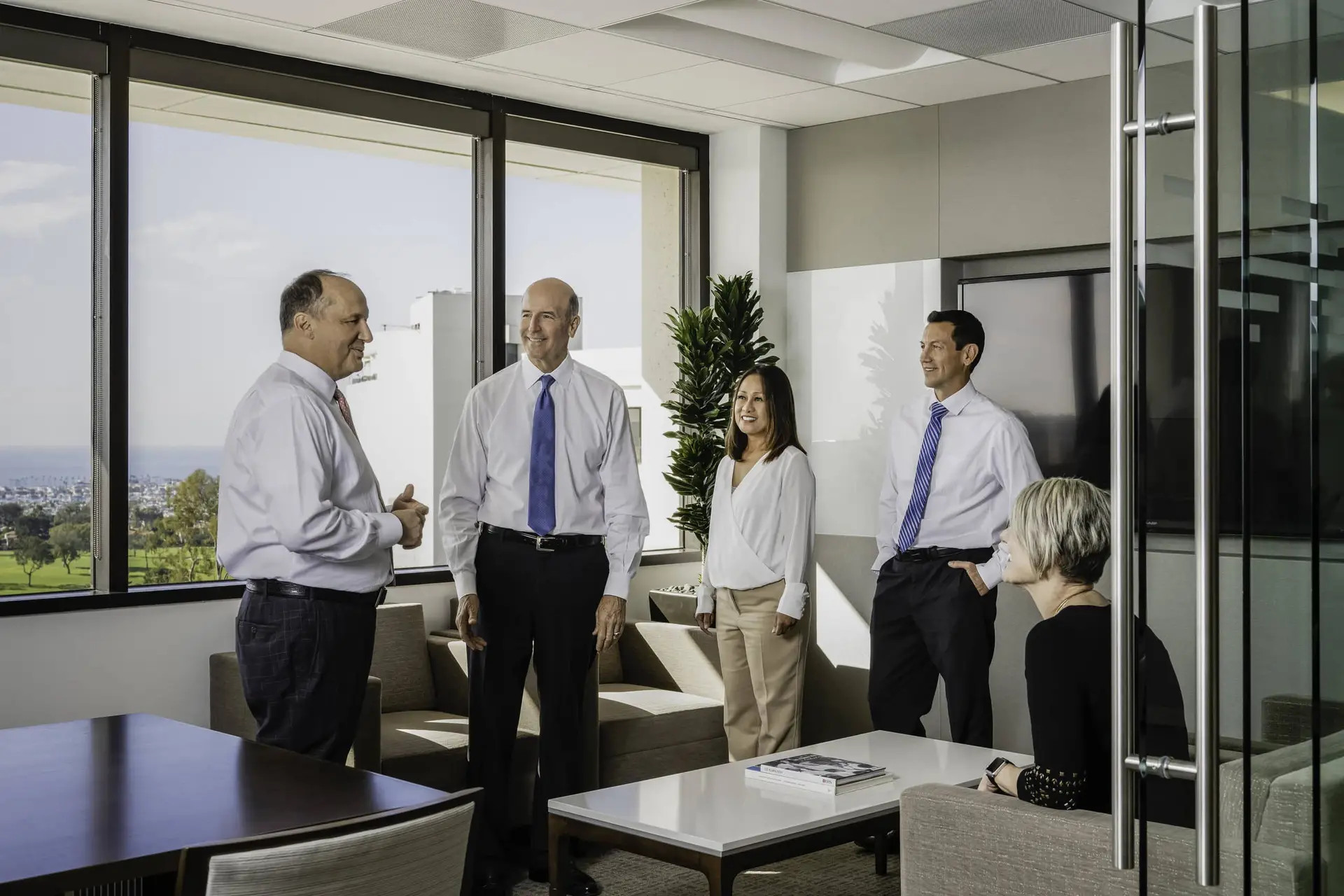 Group of professionals standing and chatting in a bright office lounge by large windows overlooking the city.