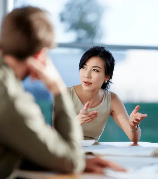 Asian woman explains a point during a business meeting, using hand gestures while a colleague listens.