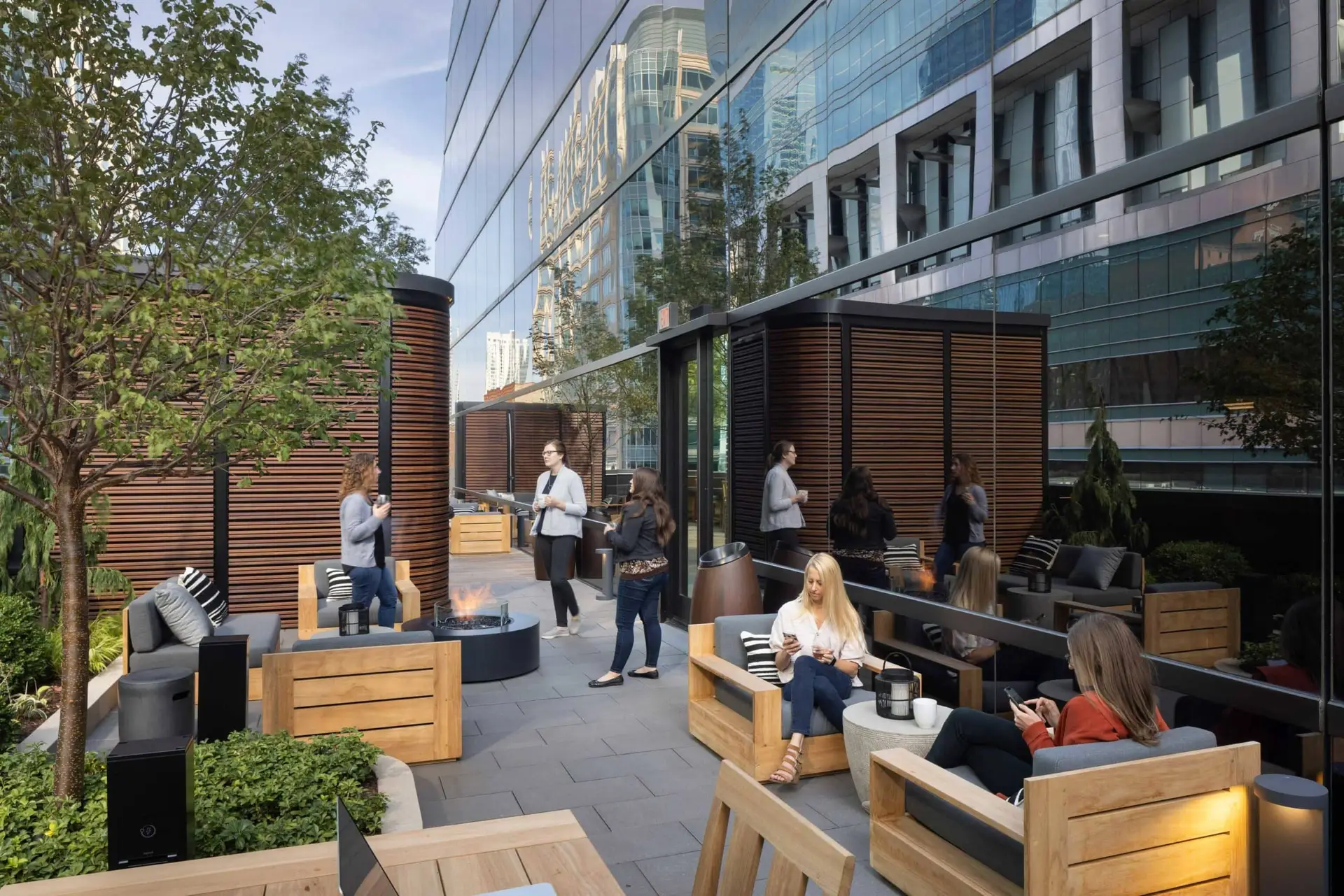 Rooftop urban lounge with wooden seating, planters, and a fire pit beside glass-walled skyscrapers.