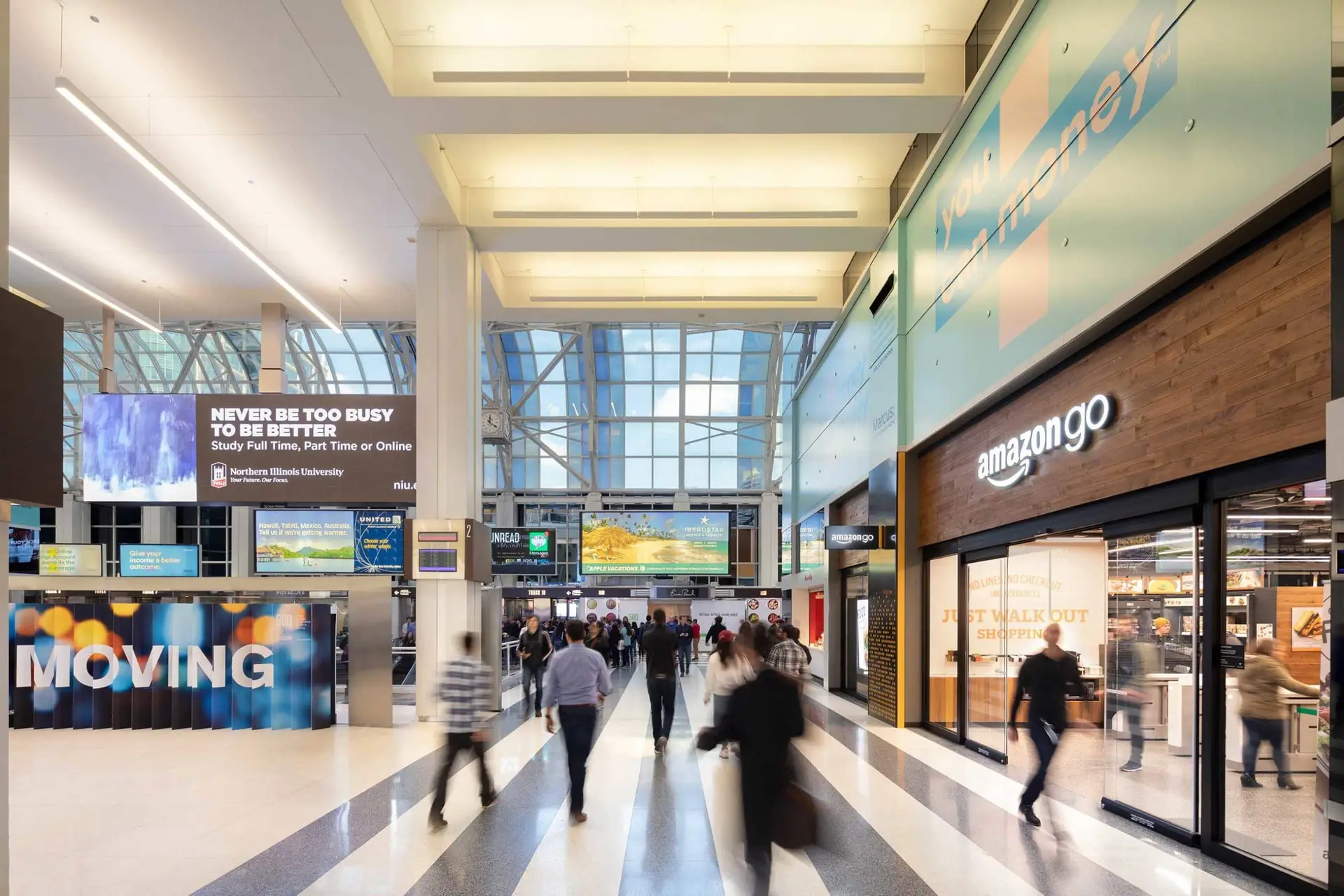 Busy airport terminal concourse with shoppers walking past an Amazon Go store on the right and a large MOVING sign on the left.
