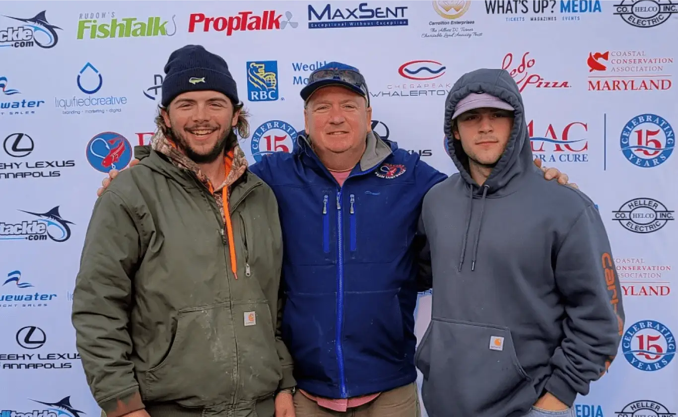 Three men posing together in front of a sponsor banner at an outdoor event.