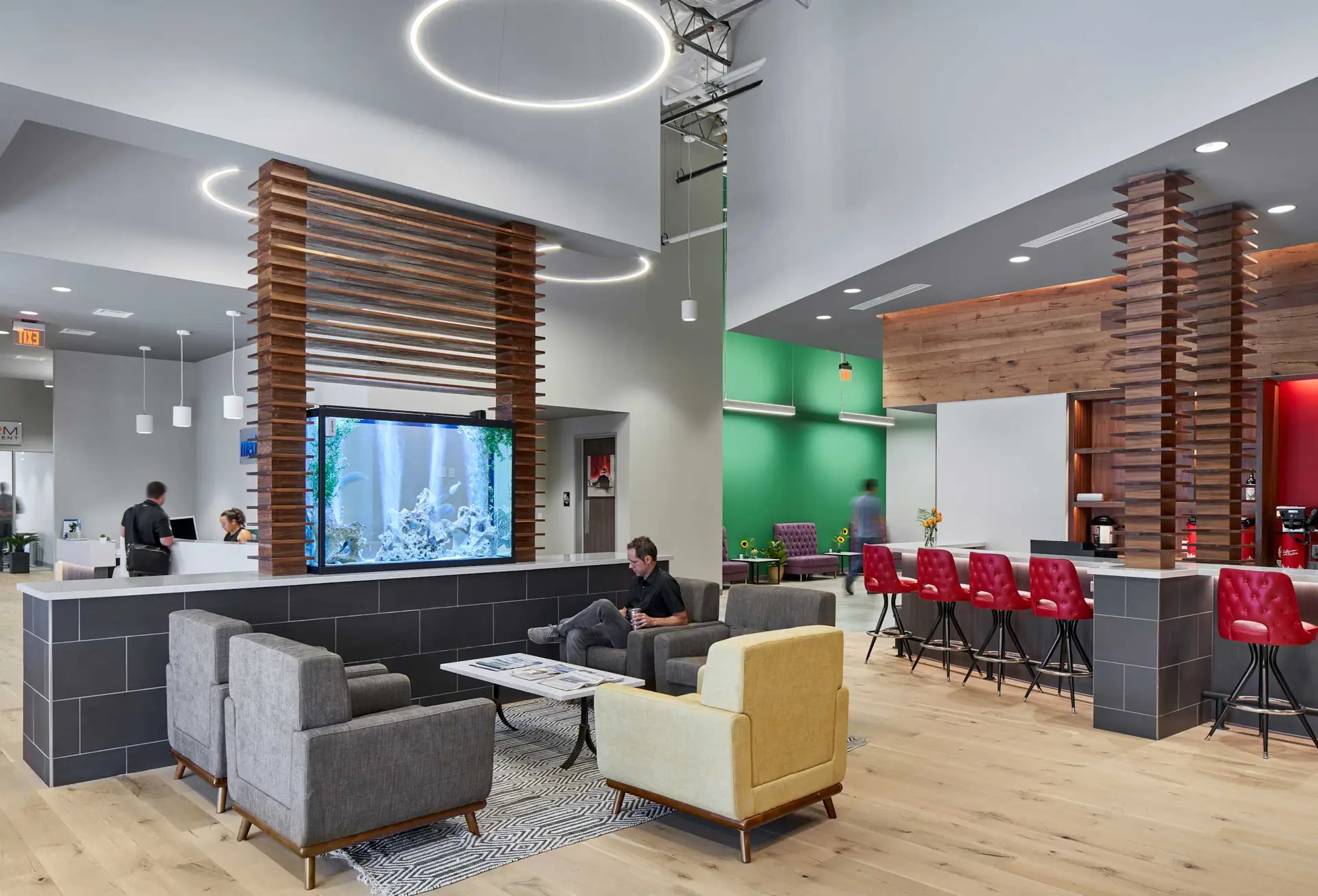 Modern office lounge with a large aquarium, circular pendant lights, wooden slat dividers, and a bar area.