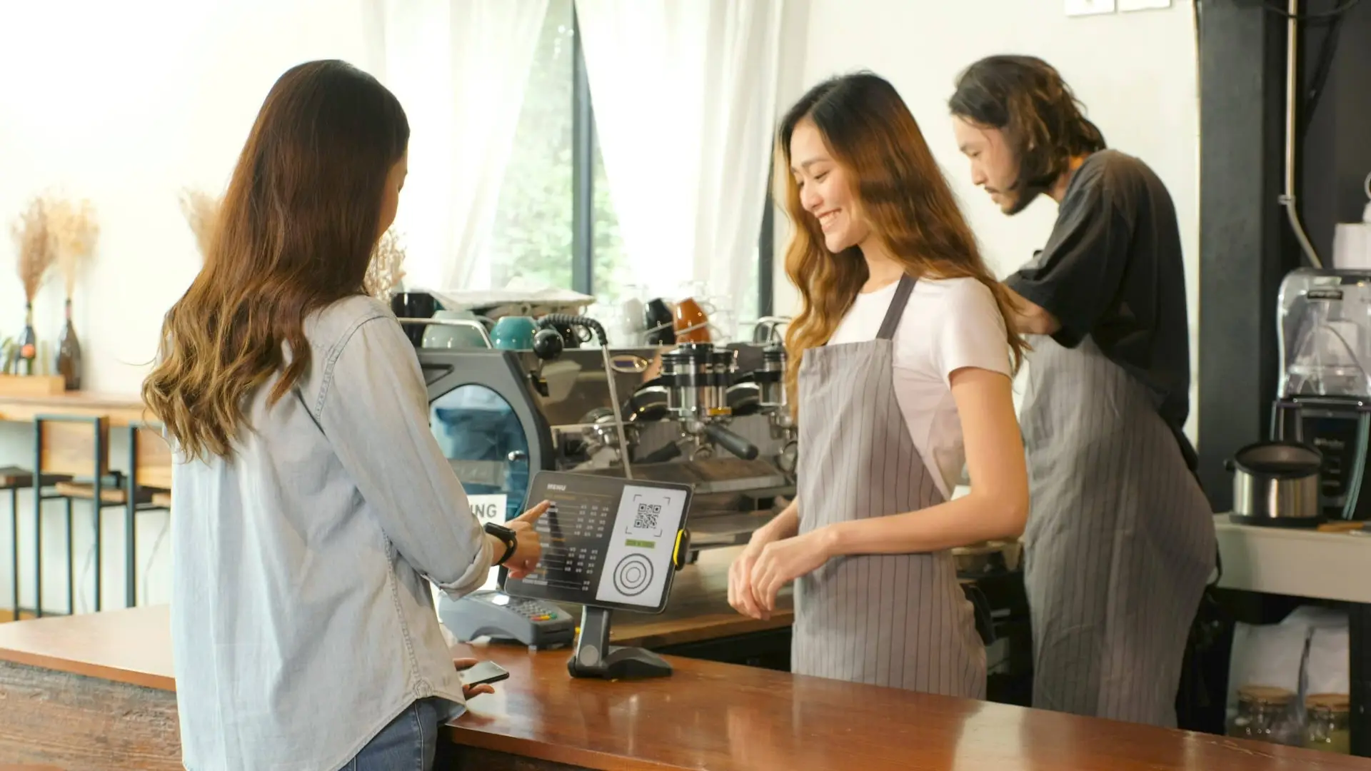 A customer pays with a tablet POS at a cafe counter while two baristas work behind the espresso machine.