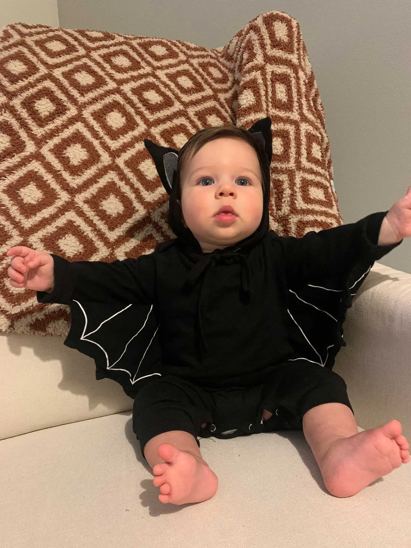 Adorable baby in a black bat costume with ears and wings, sitting on a beige couch.
