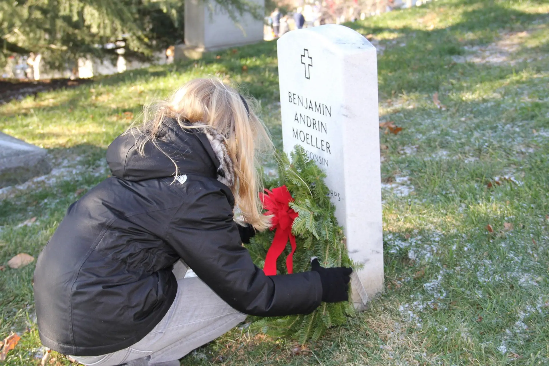 Person in a black jacket kneels beside a white gravestone, placing a green wreath with a red ribbon.