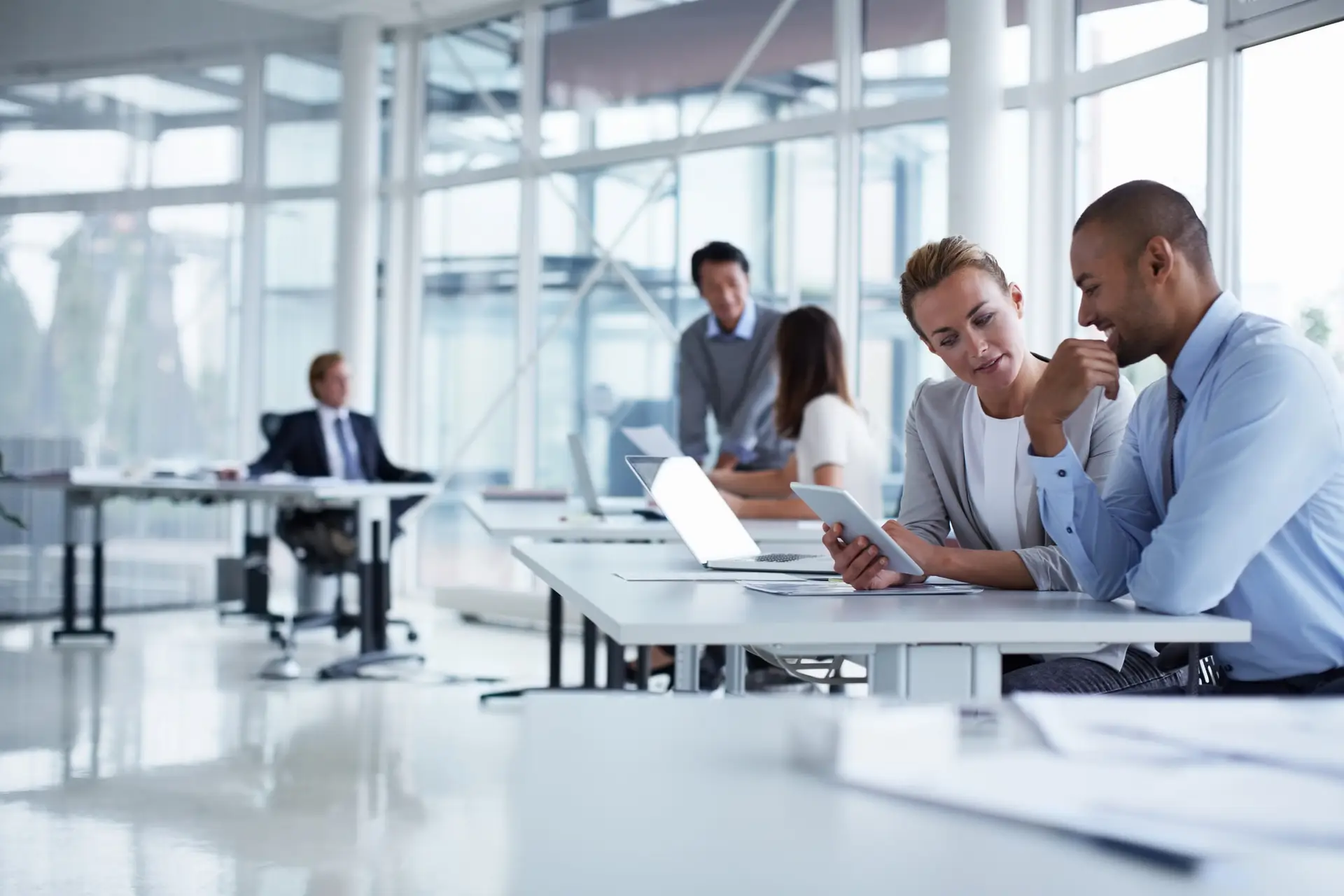 Two professionals review a tablet while seated at a white desk in a bright open-plan office.