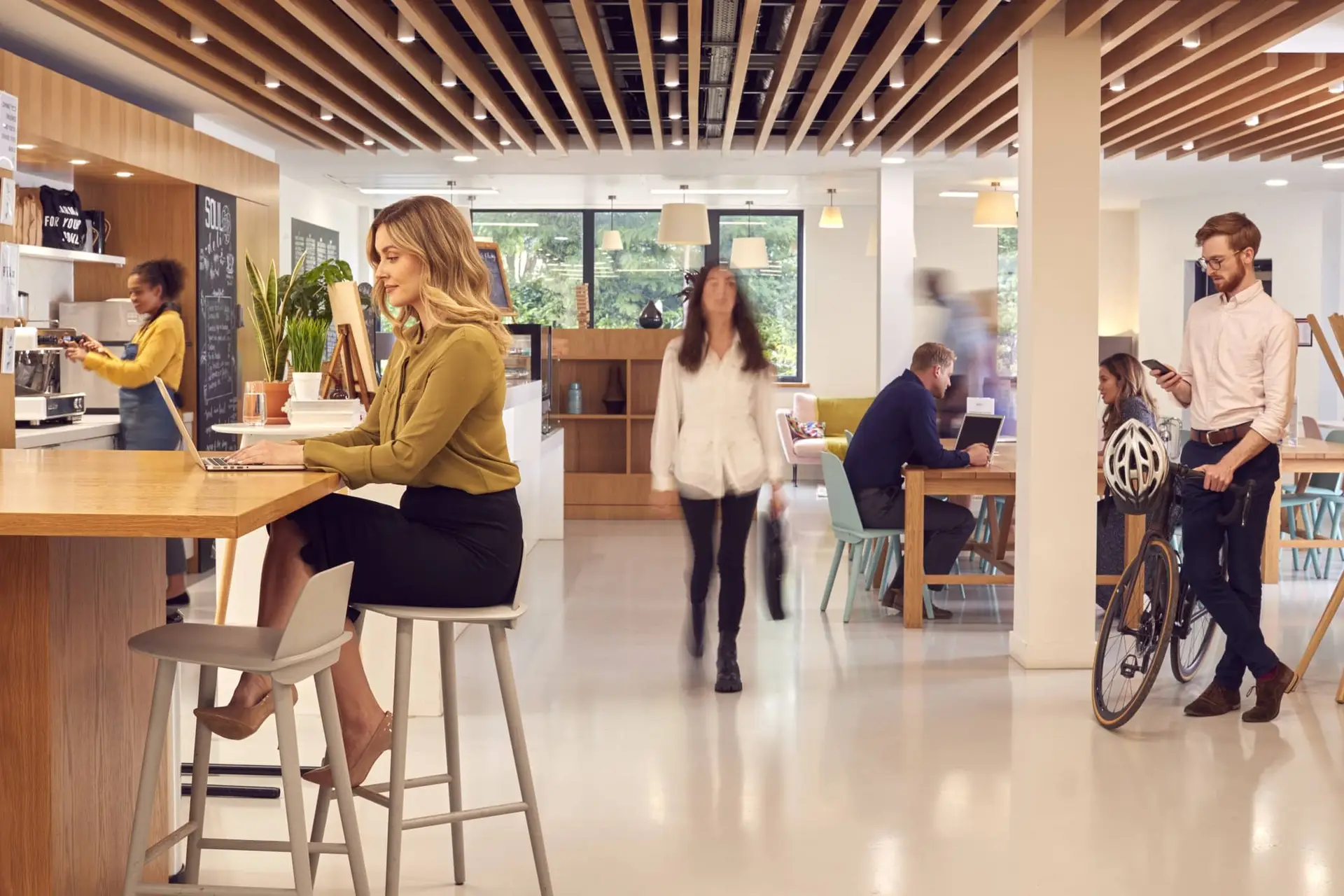 Busy modern coworking cafe with people working at a wooden counter, laptops open, and a cyclist nearby.