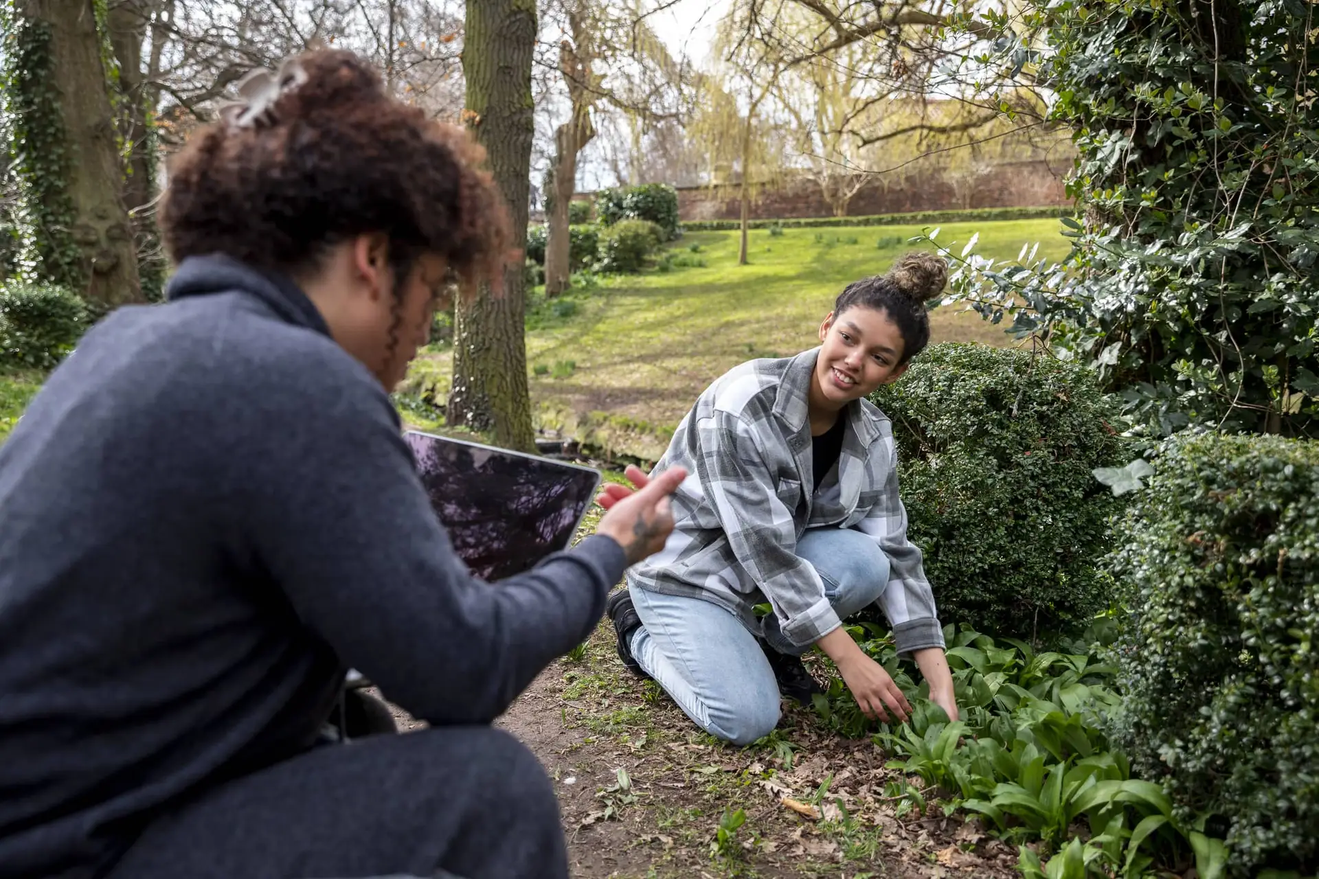 Two friends in a park garden; one kneels to tend plants while the other sits with a laptop nearby.