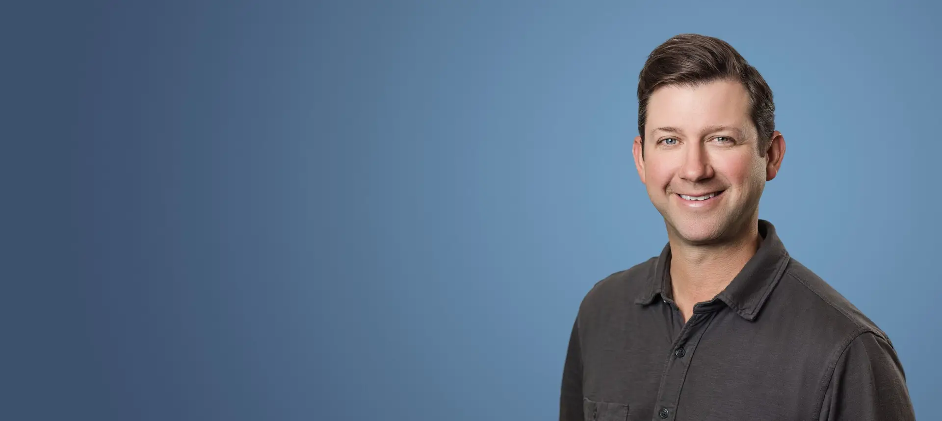 Smiling man in a dark gray button-down shirt poses against a blue gradient backdrop.