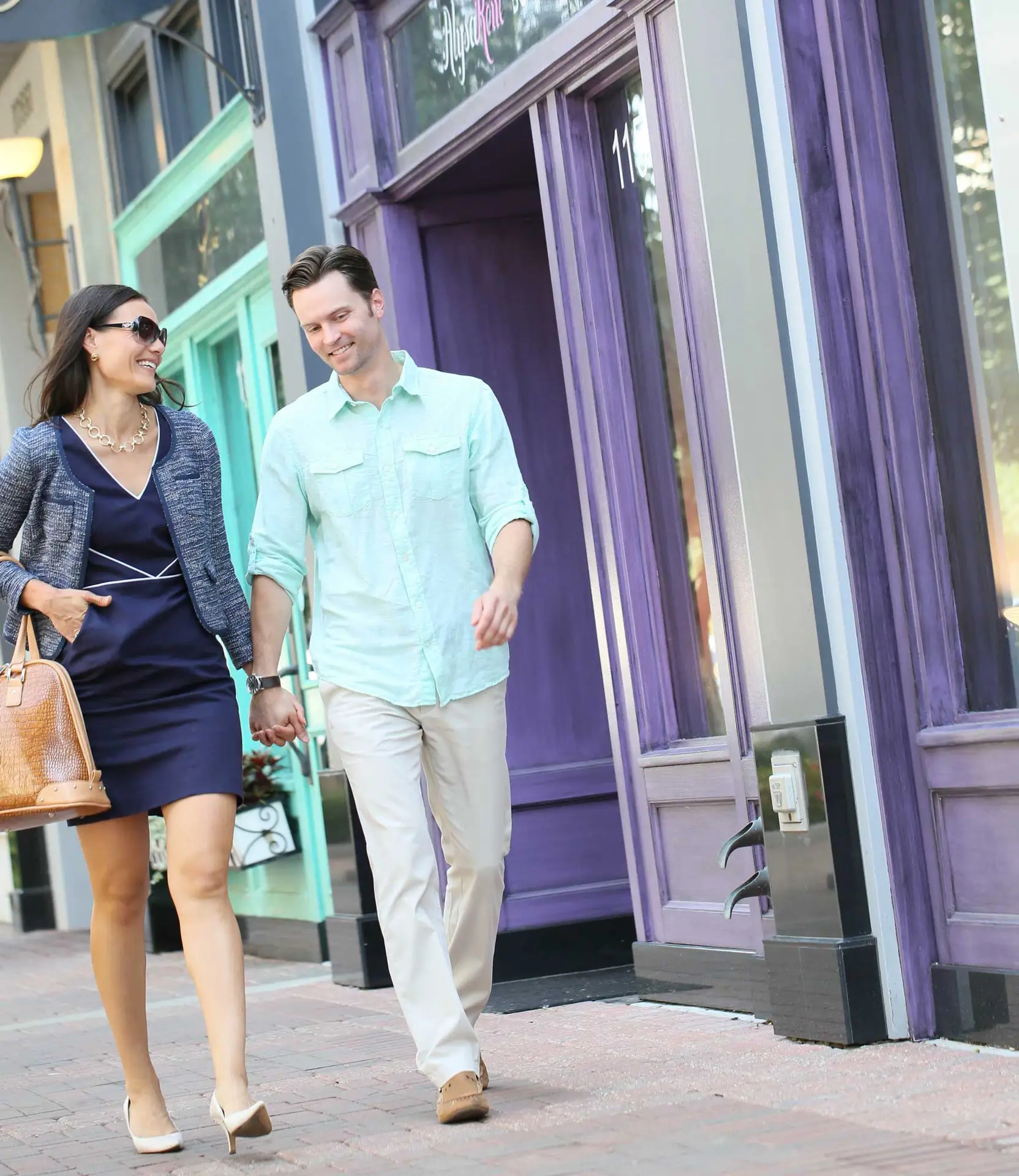 Couple walking hand in hand along a colorful storefront street, smiling at each other.