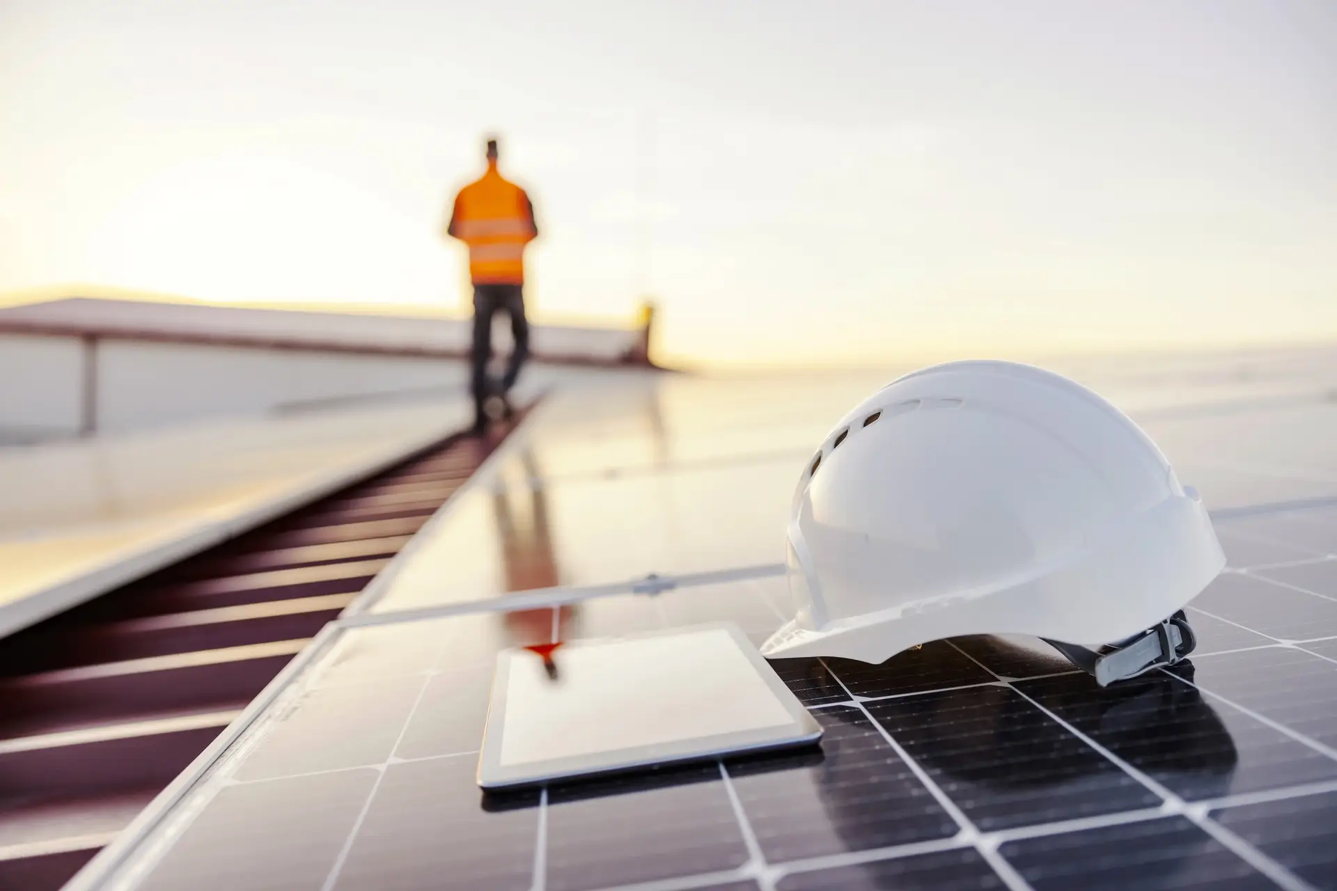 Solar rooftop with a white safety helmet in the foreground and a worker in a high-visibility vest at sunset.