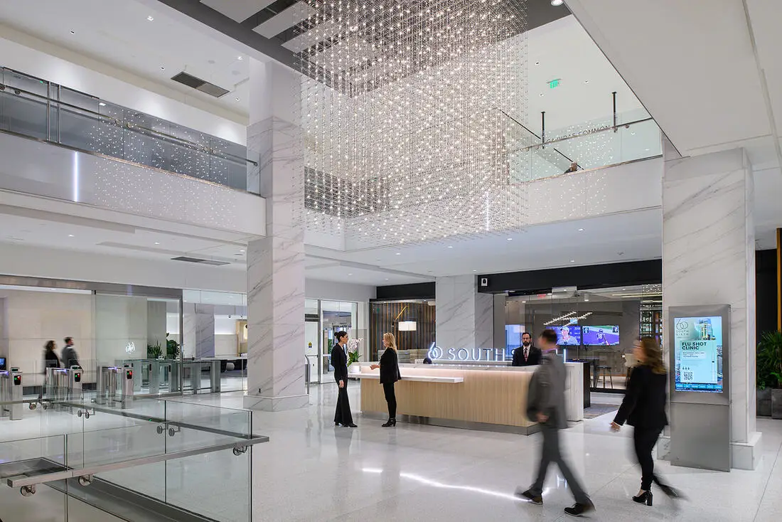 Modern corporate lobby with marble columns, glass railings, and a cascading crystal chandelier above a reception desk.