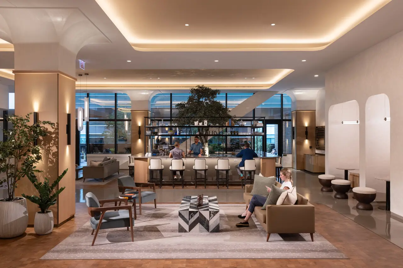 Hotel lobby with a central bar, modern seating, warm ambient lighting, and large windows.