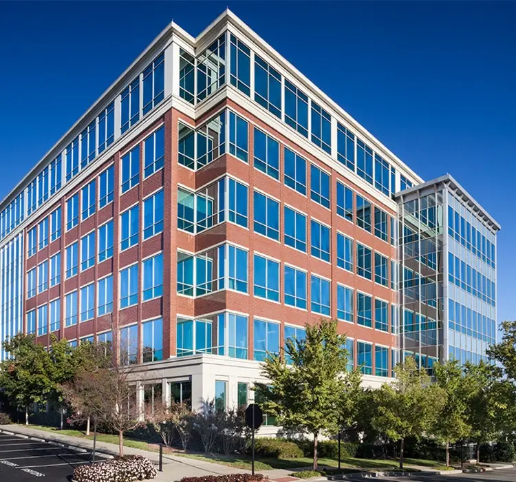 Modern brick-and-glass office building with numerous windows and surrounding trees against a clear blue sky.