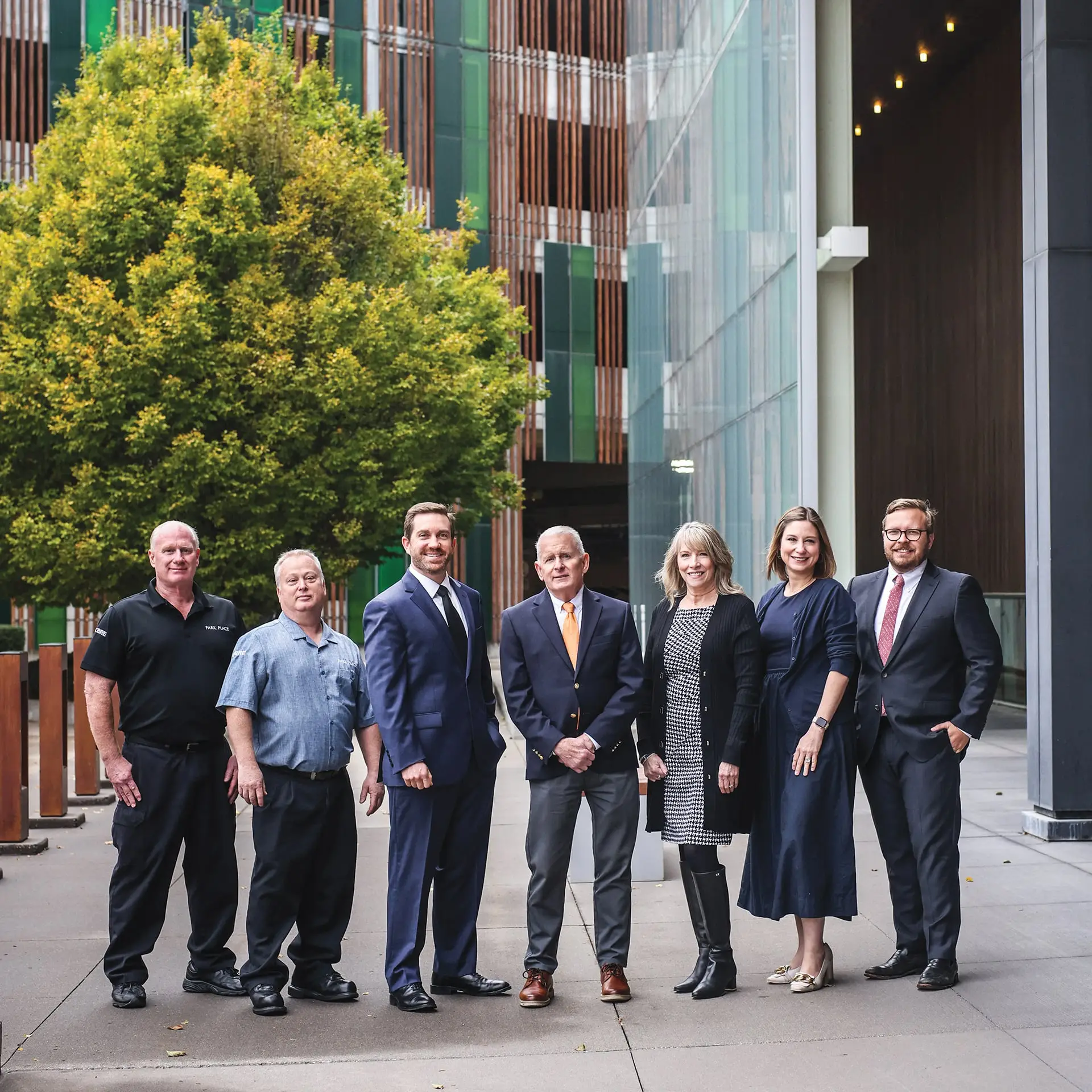 Seven people stand smiling on a city sidewalk, lined up beside a large leafy tree.