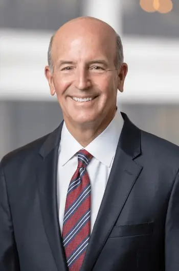 Bald man smiles, wearing dark suit and striped tie, against blurred background.