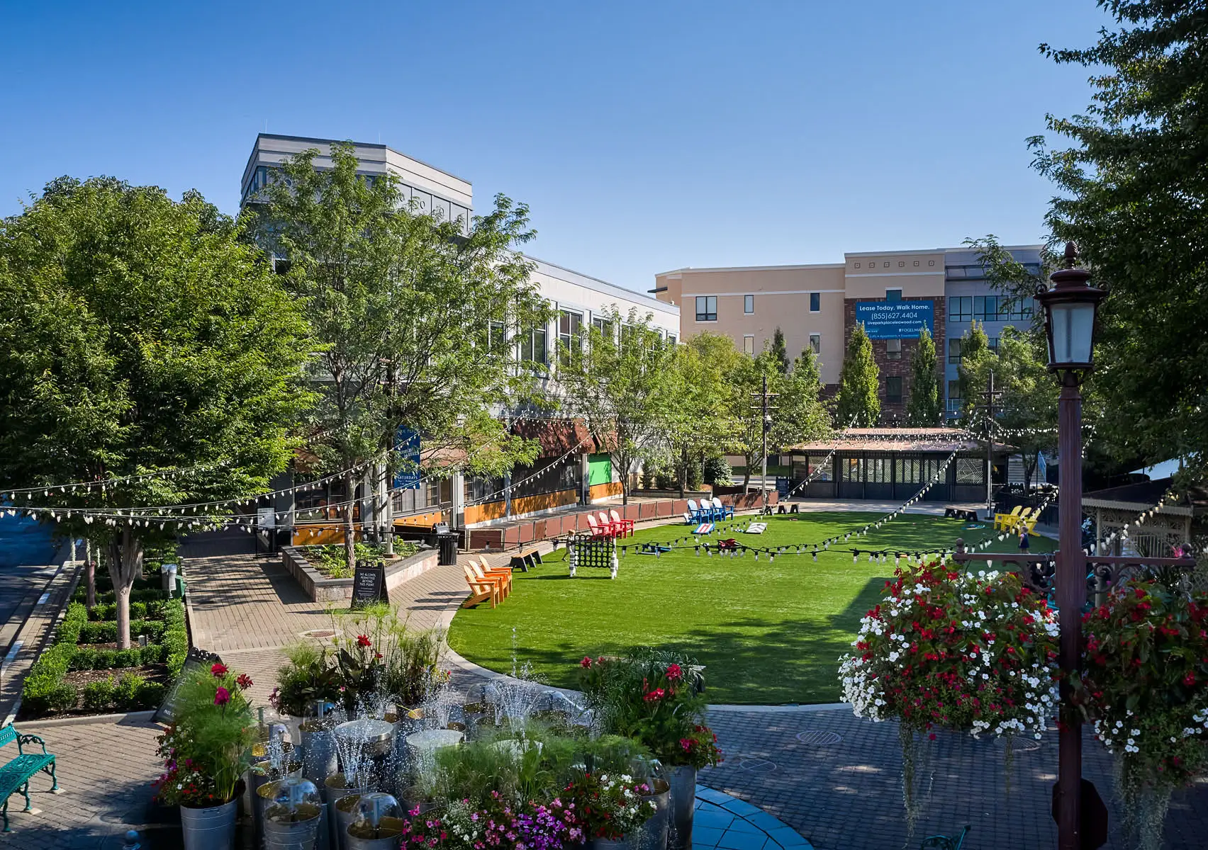 Campus courtyard with green lawn, string lights, trees, and outdoor seating against modern buildings.