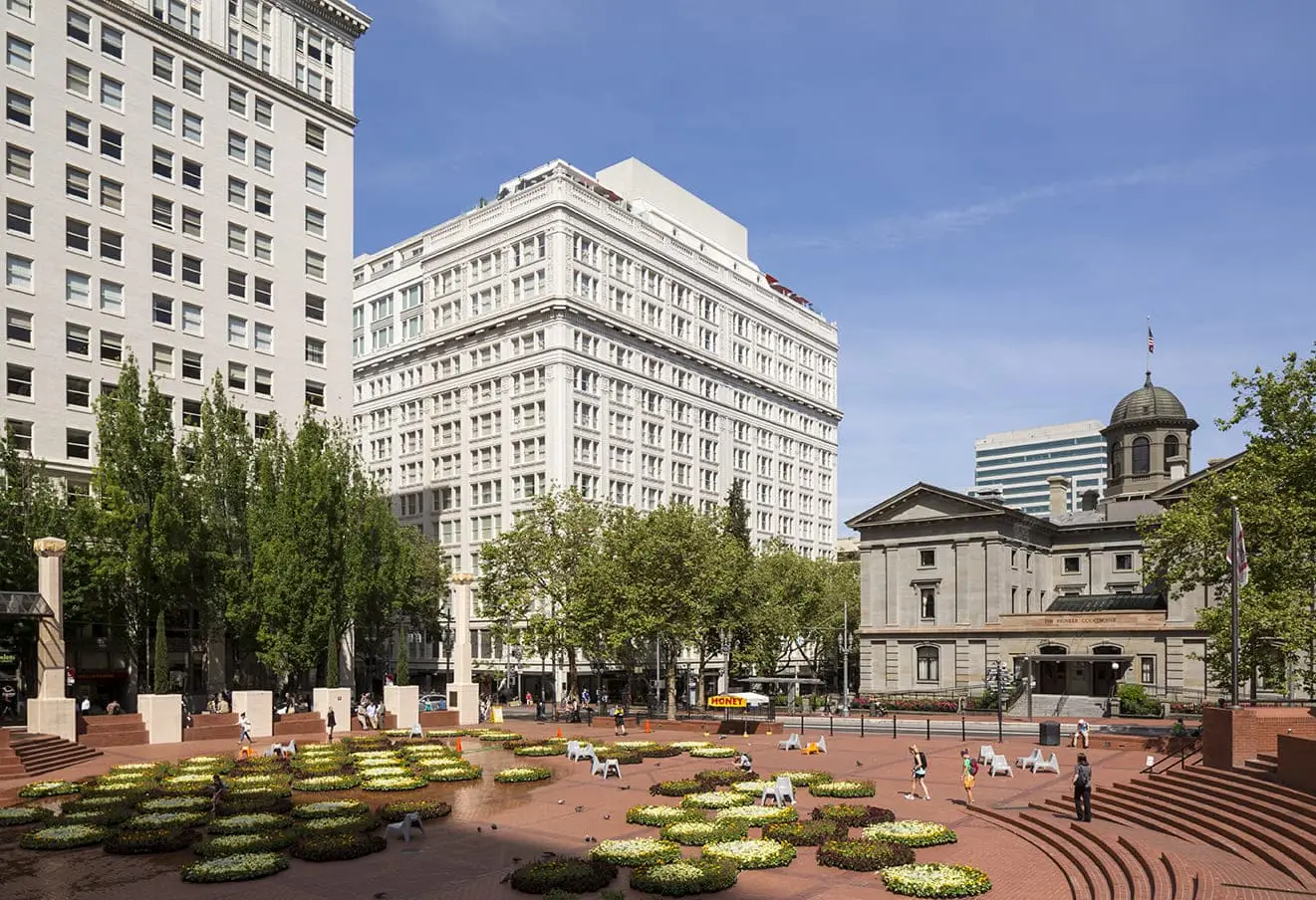 Urban plaza with circular flower beds, brick pavement, surrounding tall white buildings and a domed courthouse in the background.
