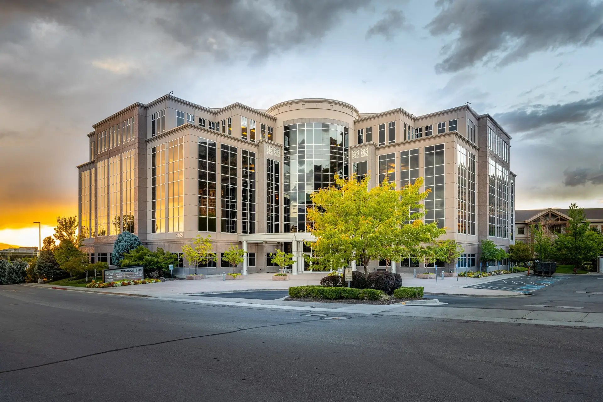 Modern office building with a curved glass atrium, tall glass façades, and a landscaped plaza at sunset.