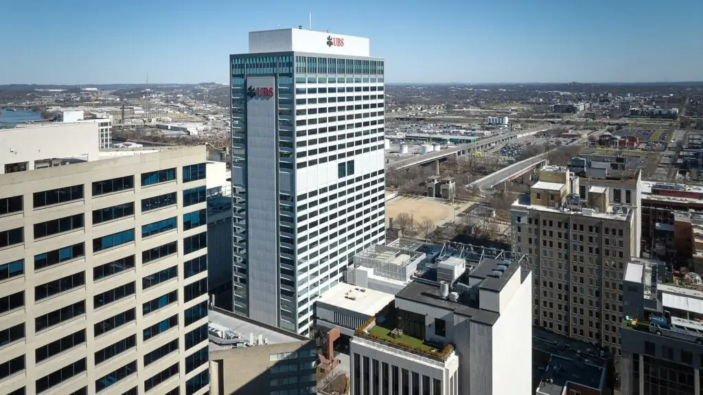 Aerial view of a city with a tall UBS office tower surrounded by low-rise buildings and streets beneath a clear blue sky.