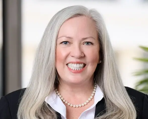 Professional portrait of a smiling woman with long silver hair, pearl necklace, white blouse, and black blazer.