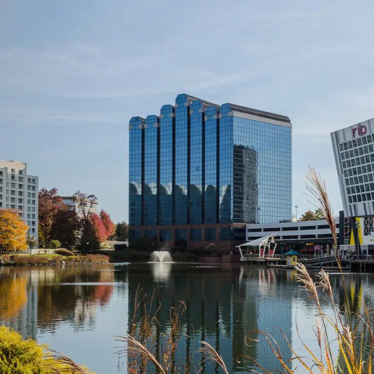 Glass skyscraper reflects in calm lake; autumn trees and modern buildings surround.