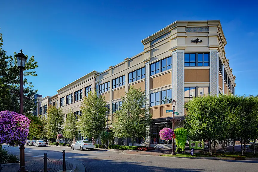 Modern beige brick office building with large glass windows, tree-lined street, and purple flowering planters.