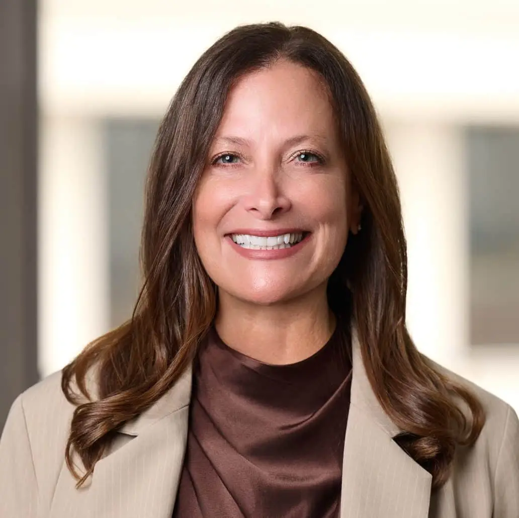 Smiling woman with long brown hair in a beige blazer and brown blouse in an office setting.