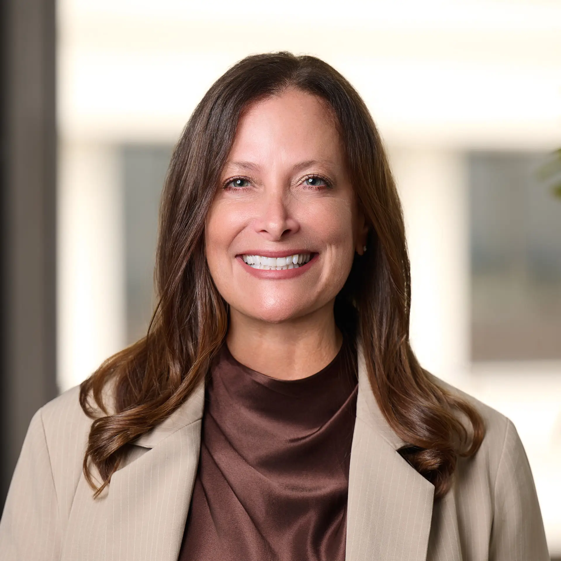 Smiling woman in a beige blazer and brown blouse in a bright office setting.
