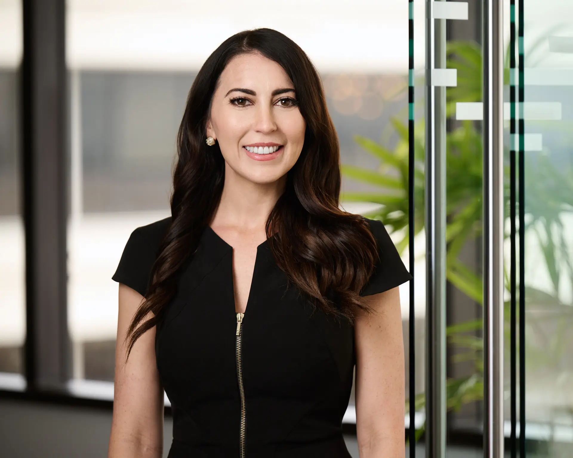 Smiling woman in a black dress stands in a modern office with glass doors and plants.