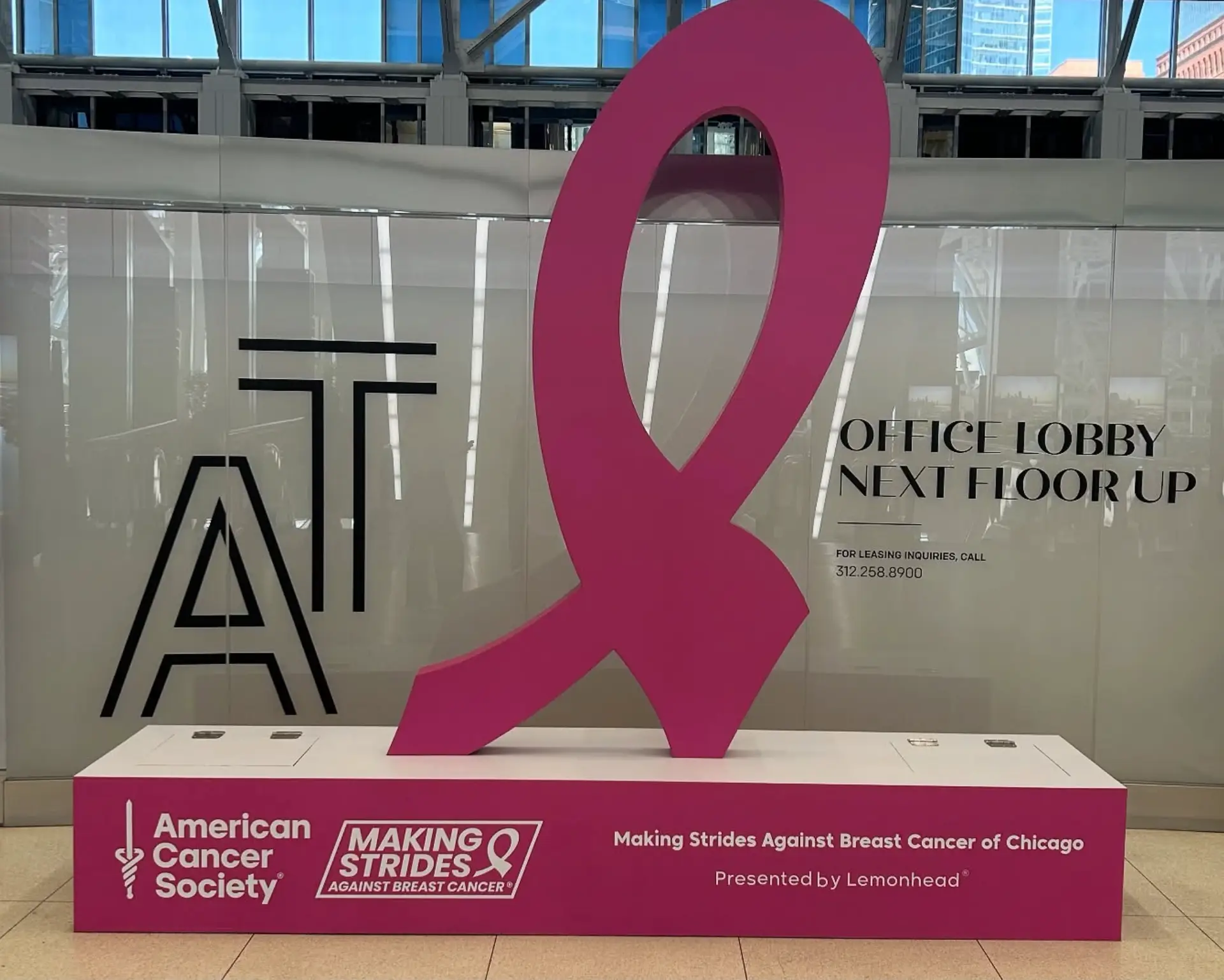 Large pink breast cancer awareness ribbon sculpture on a magenta pedestal in an office lobby.