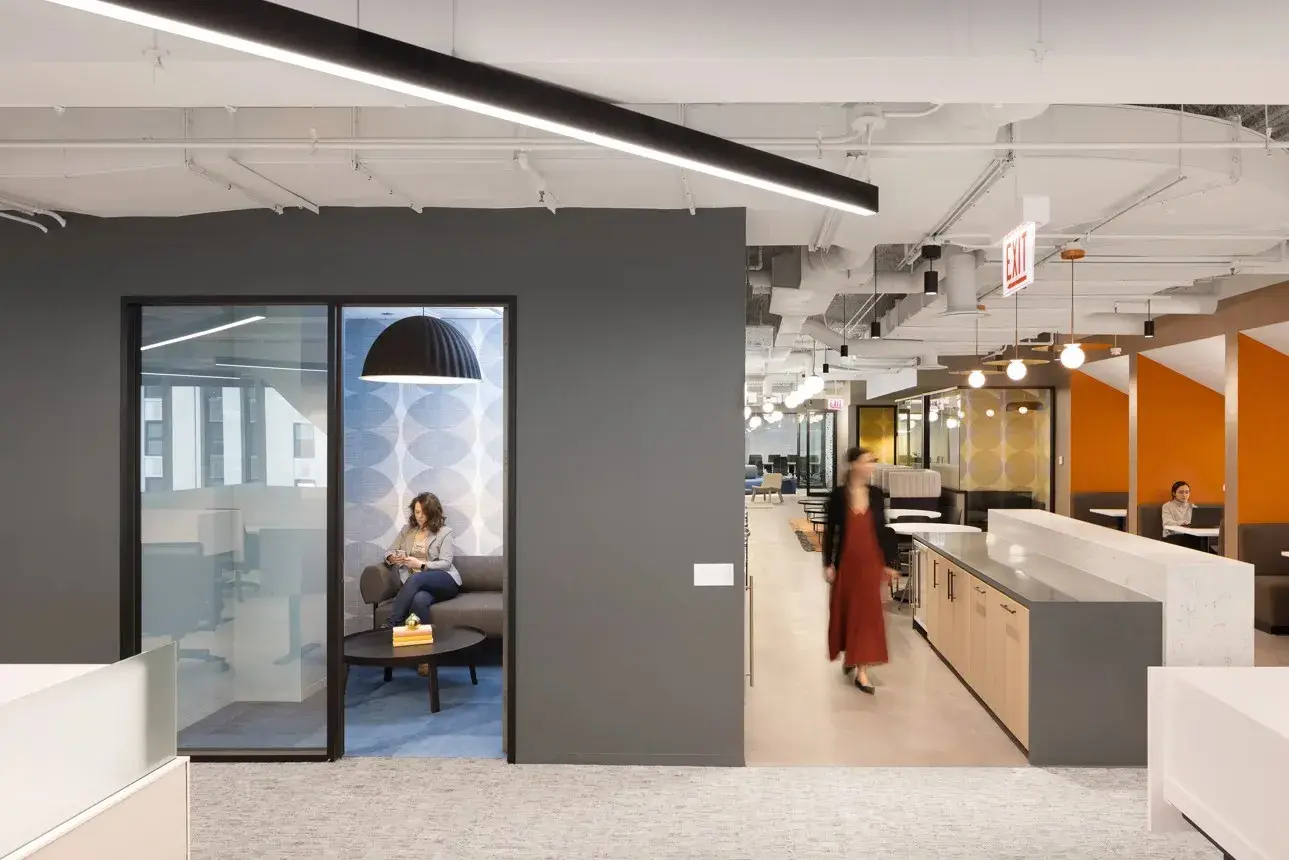 Modern open-plan office with a glass-walled meeting room, a person walking past a kitchen island, and orange accent walls.