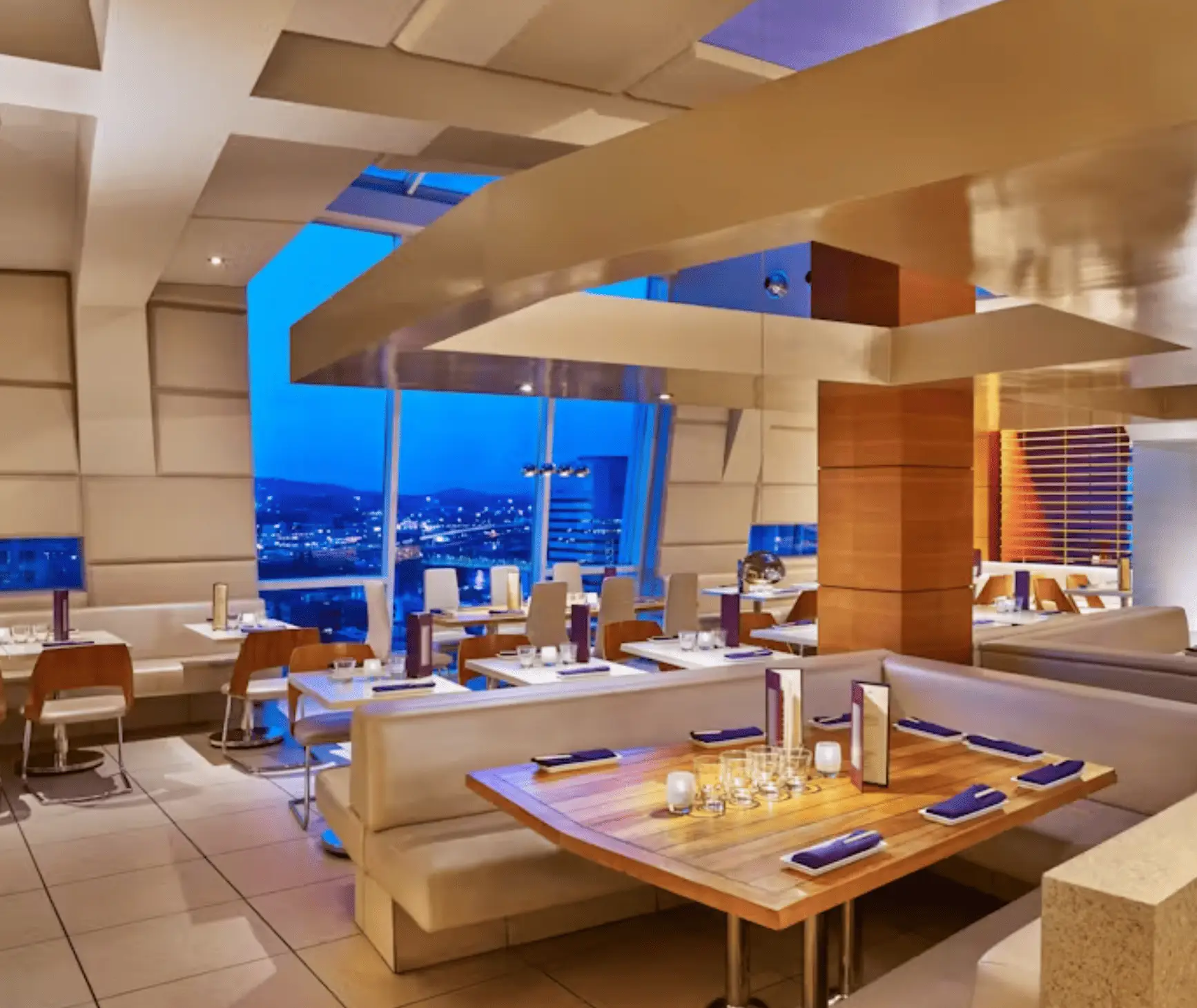 Modern restaurant interior with beige seating, wooden tables, and blue-lit windows overlooking a city at dusk.