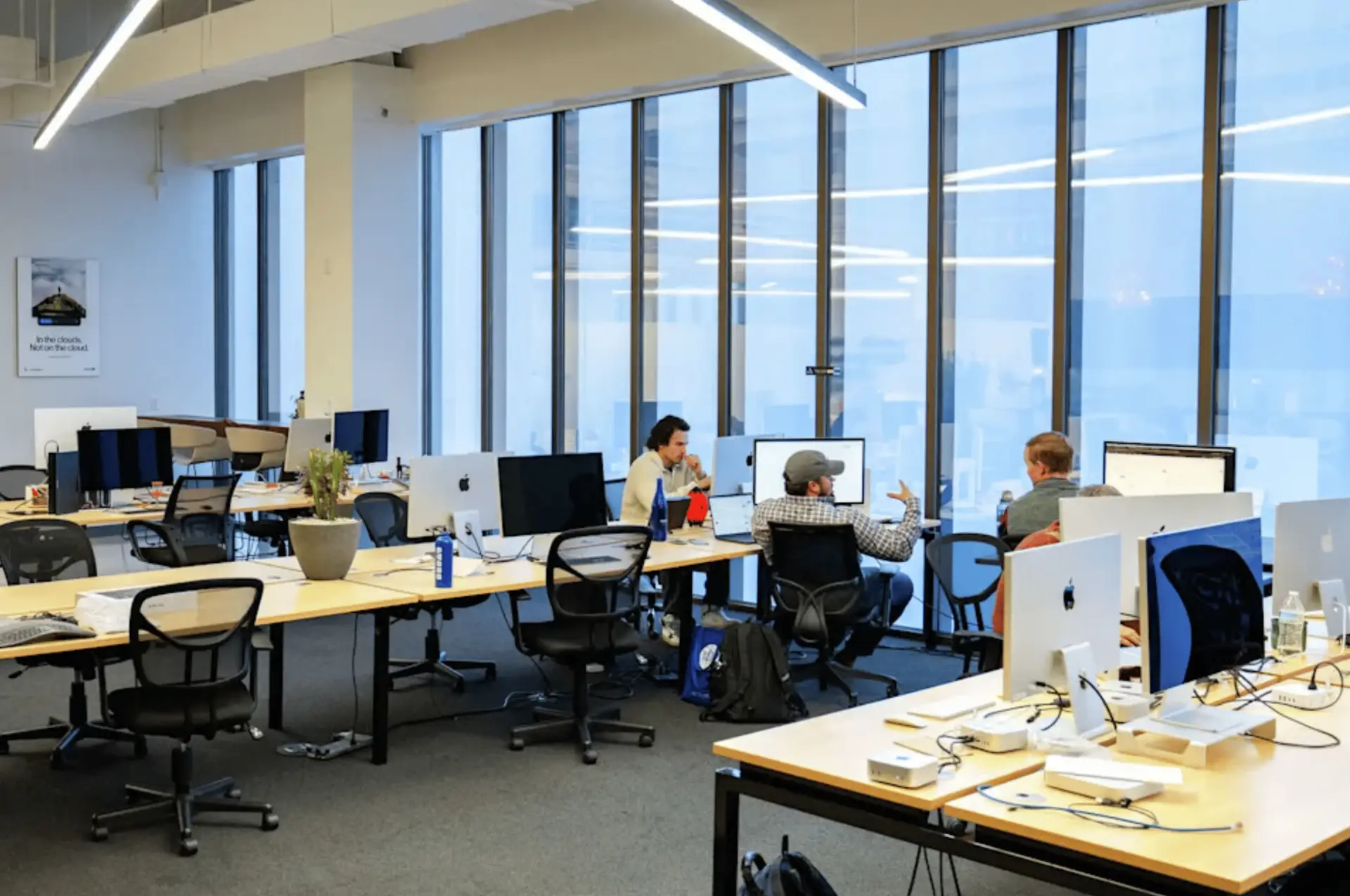 Open-plan coworking office with rows of desks, computer monitors, and tall glass windows.